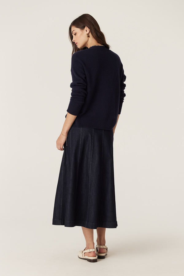 Model wearing a dark indigo Burke denim skirt with gored panels and contrast stitching, paired with a navy sweater.