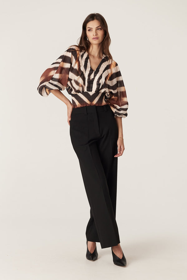 Woman wearing a relaxed fit Sasha Blouse with brown zebra print, V-neck, batwing sleeves, and shirred waist, paired with black trousers.
