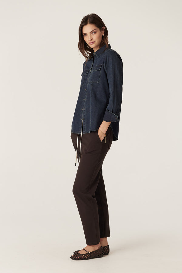 Woman wearing an indigo Dallas denim shirt with patch pockets and cream press studs, paired with dark pants.