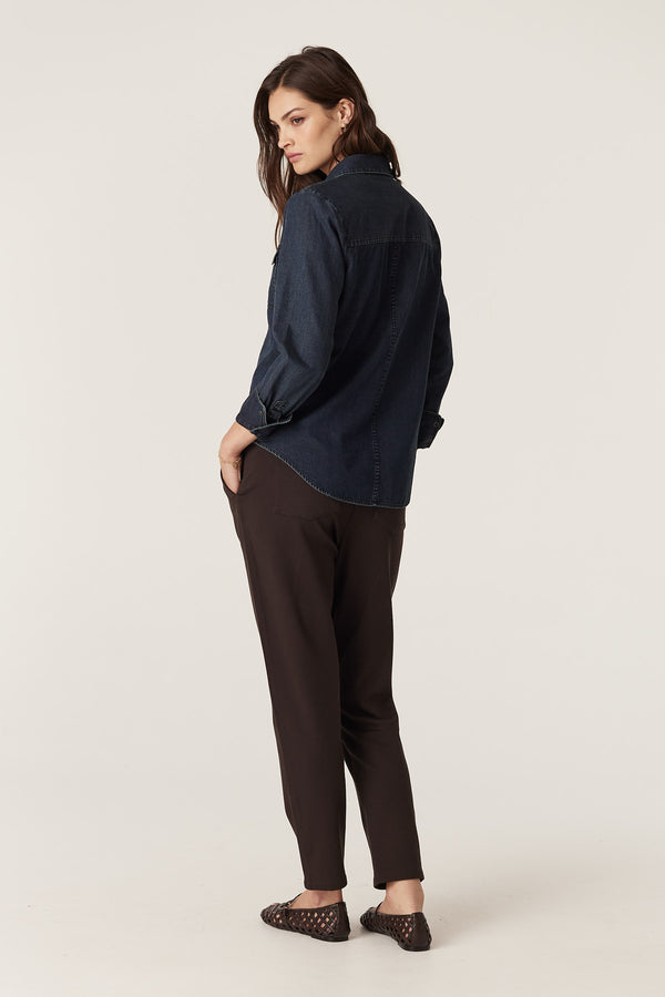 Woman in indigo Dallas denim shirt with patch pockets and dark pants, viewed from behind.