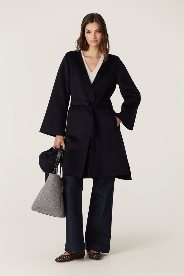 Woman in navy Lawson wool coat with flared sleeves, high-low hem, and matching belt, holding a grey bag.