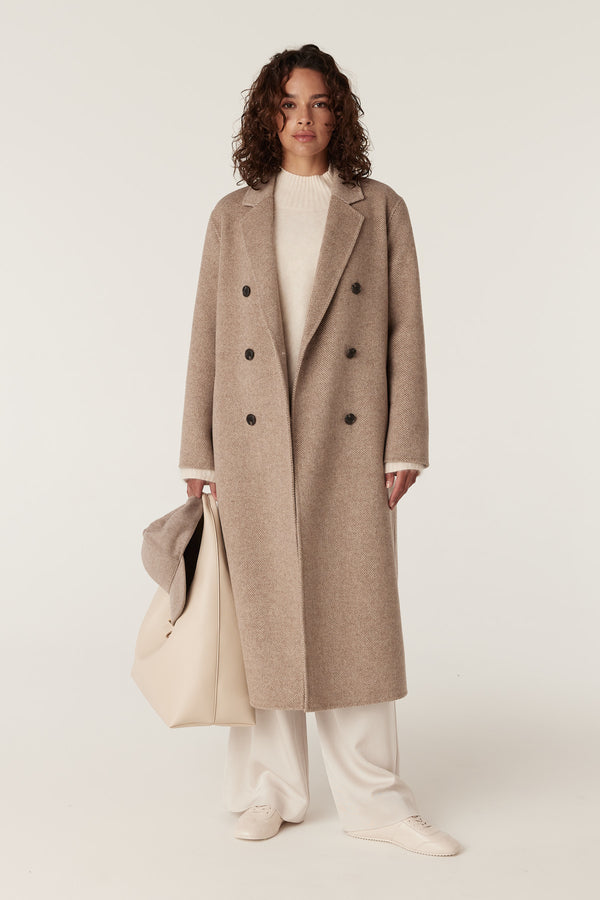 Woman in a fawn Ainsley wool blend coat, cream sweater, white pants, holding a matching tote bag.