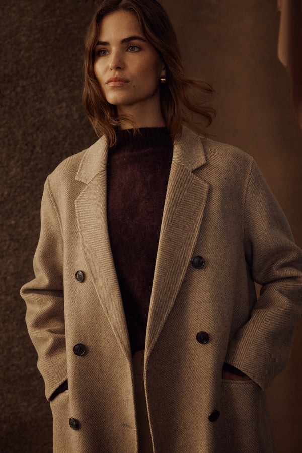 Woman wearing a fawn Ainsley double-breasted coat with wide lapels and dark buttons over a burgundy sweater.