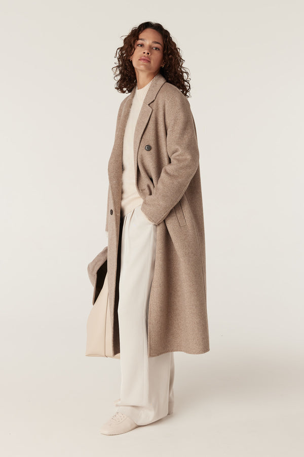 Woman in a fawn Ainsley wool coat, cream sweater, white pants, and sneakers, holding a light tote bag.