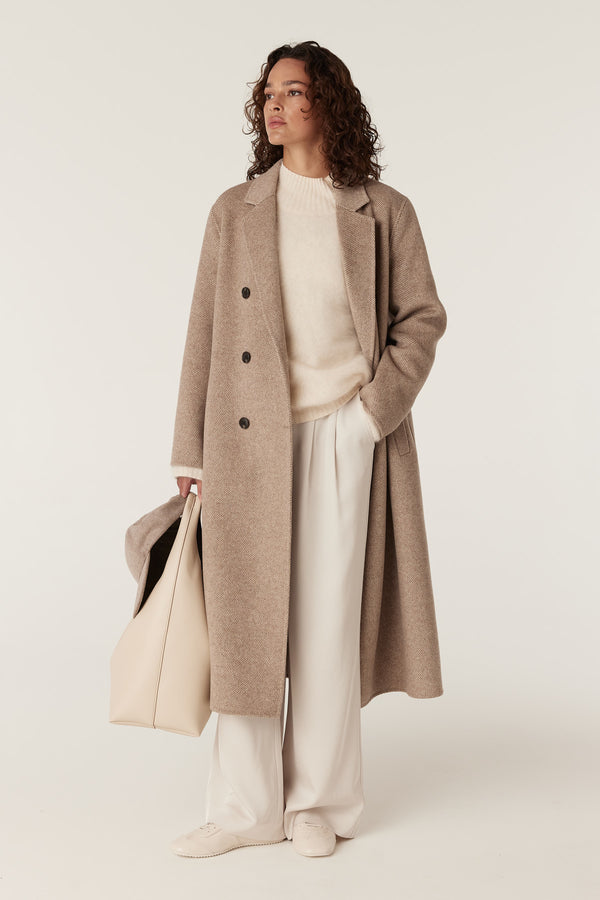 Woman wearing a fawn Ainsley wool coat, cream sweater, wide-leg pants, and holding a matching tote bag.