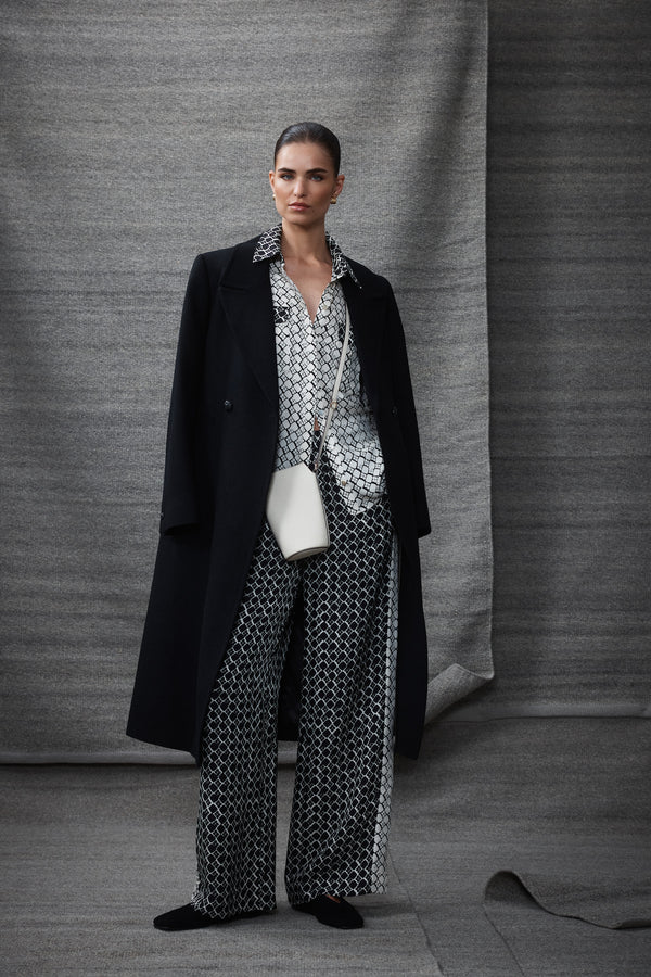 Woman wearing a black Montgomery wool coat, white patterned shirt, black patterned pants, and white shoulder bag.