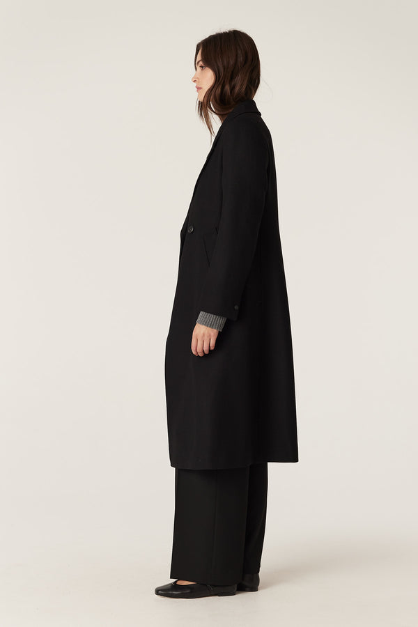 Woman in a black wool twill Montgomery coat with revere collar, double-breasted front, and functional pockets.