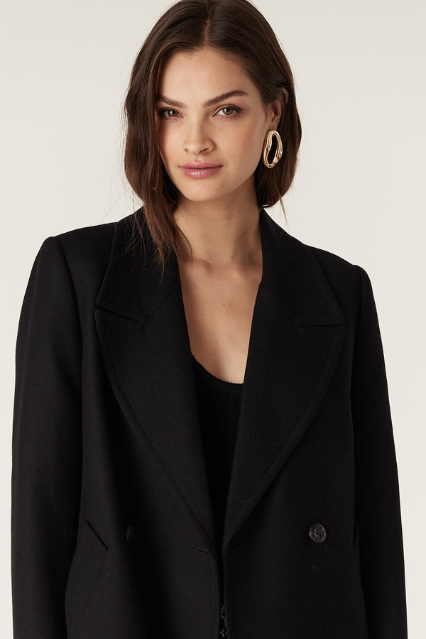 Woman wearing a black wool twill double-breasted Montgomery coat with revere collar and metal buttons.