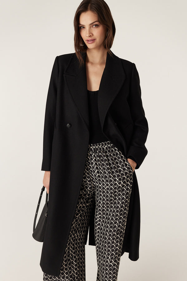 Woman wearing a black wool twill double-breasted Montgomery coat with revere collar and patterned trousers.