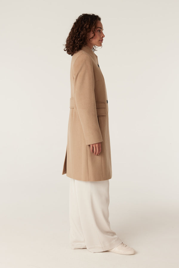 Woman wearing a camel Eton wool coat with a stand collar and side pockets.