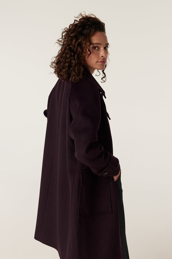 Woman in a dark port purple Oxford wool coat with large front pockets, looking over her shoulder.