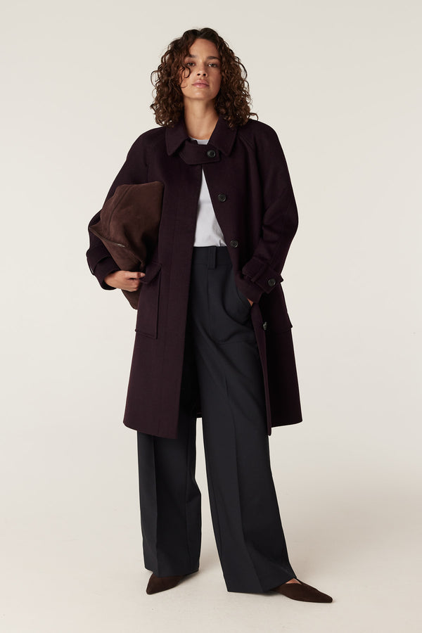 Woman in a port-colored Oxford wool coat with a classic collar, button front, and wide sleeves, holding a brown clutch.