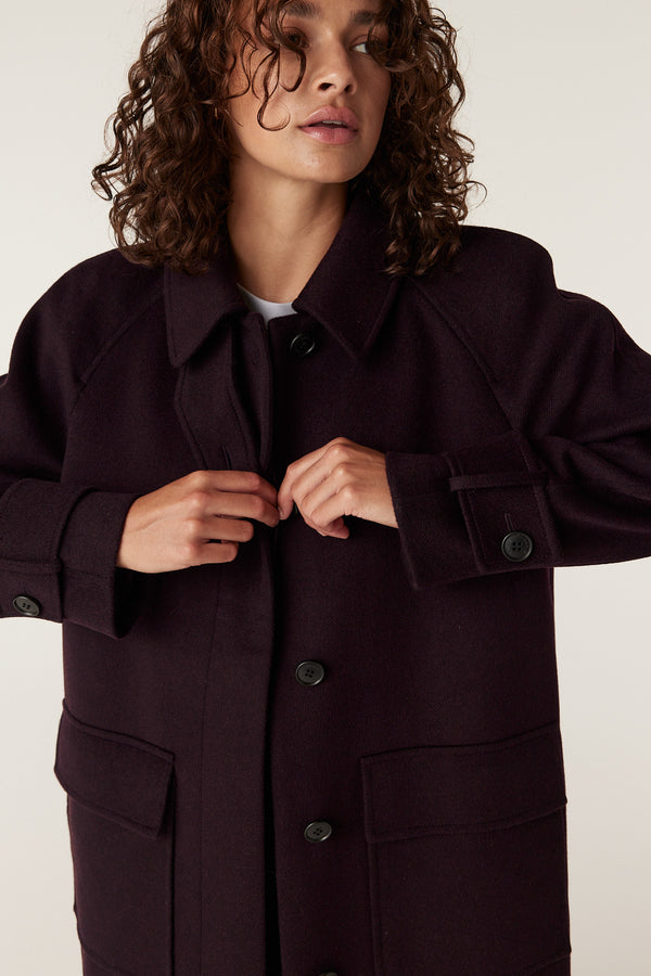 Woman fastening a dark port-colored Oxford coat with large black buttons, featuring a classic collar and front pockets.
