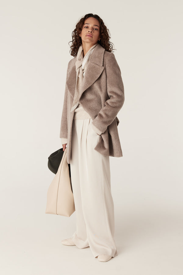 Woman in a fawn Sinclair coat, cream sweater, wide-leg pants, holding a beige tote and black hat.
