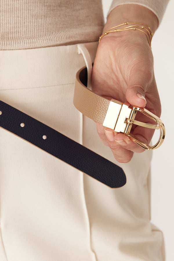 Hand holding a genuine leather reversible tan and navy belt with a gold buckle.