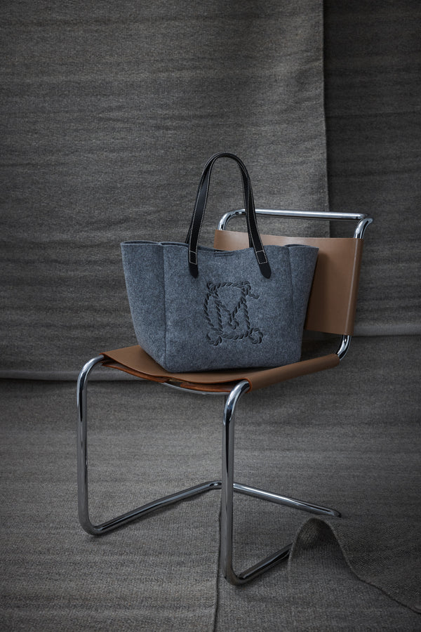 Charcoal marle wool blend felt tote with black leather handles and embroidered logo, on a modern chair.