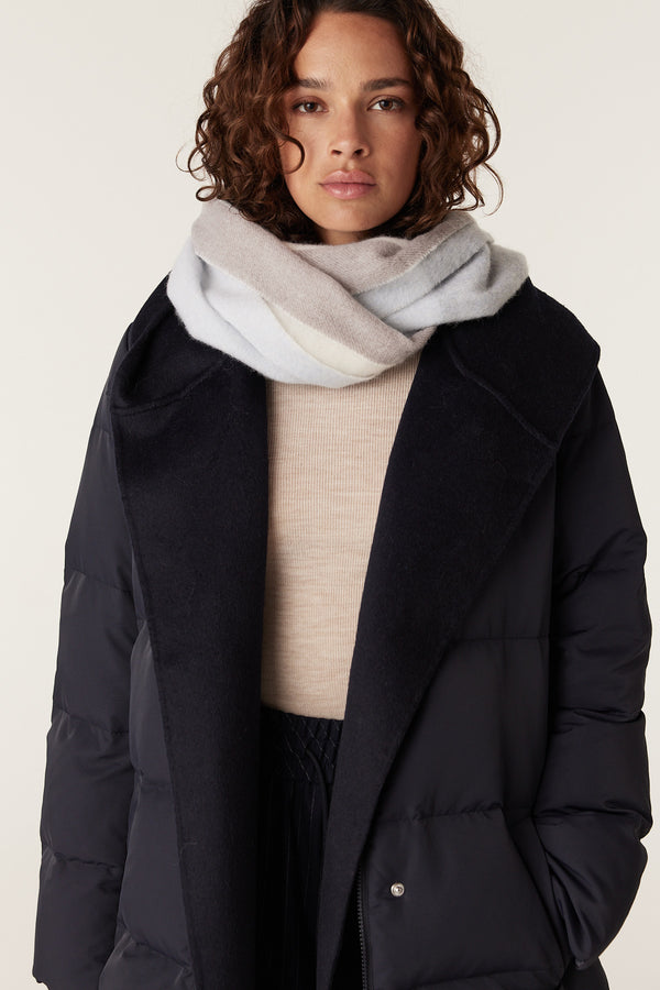 Woman wearing a soft, striped wool scarf in light blue, beige, and white, over a navy puffer coat.