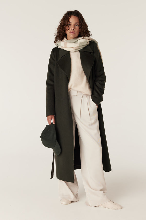 Model wearing a sage check pure wool scarf, draped over a dark green coat and light trousers.