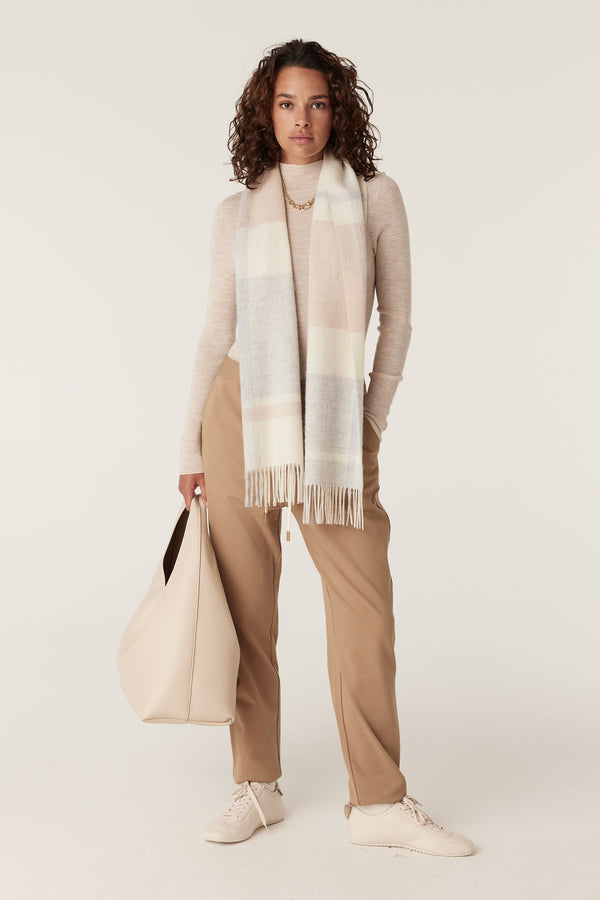 Woman wearing a blush check pure wool scarf with fringe, a beige top, and tan pants, holding a cream bag.