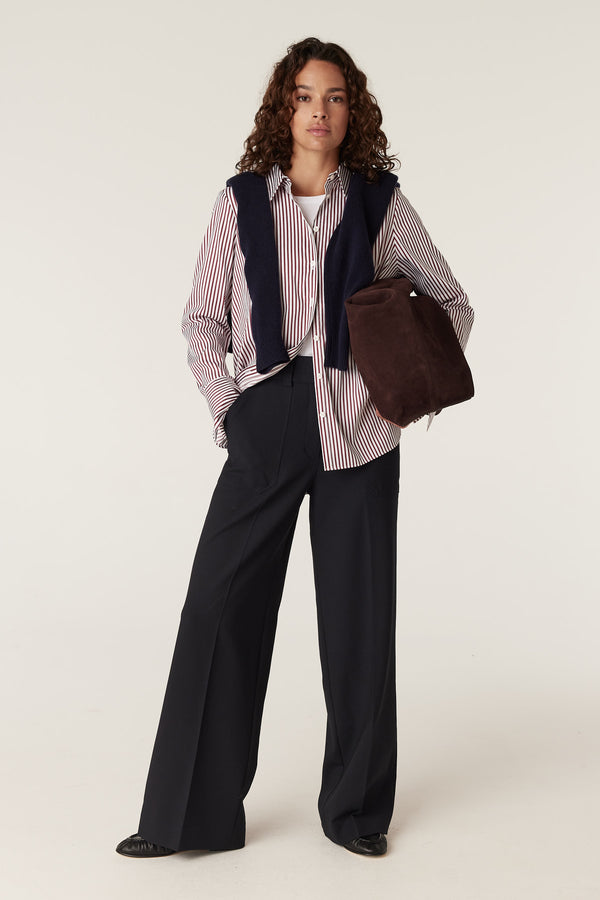 Woman wearing a red stripe Henri shirt, navy sweater vest, black wide-leg pants, and holding a brown suede bag.