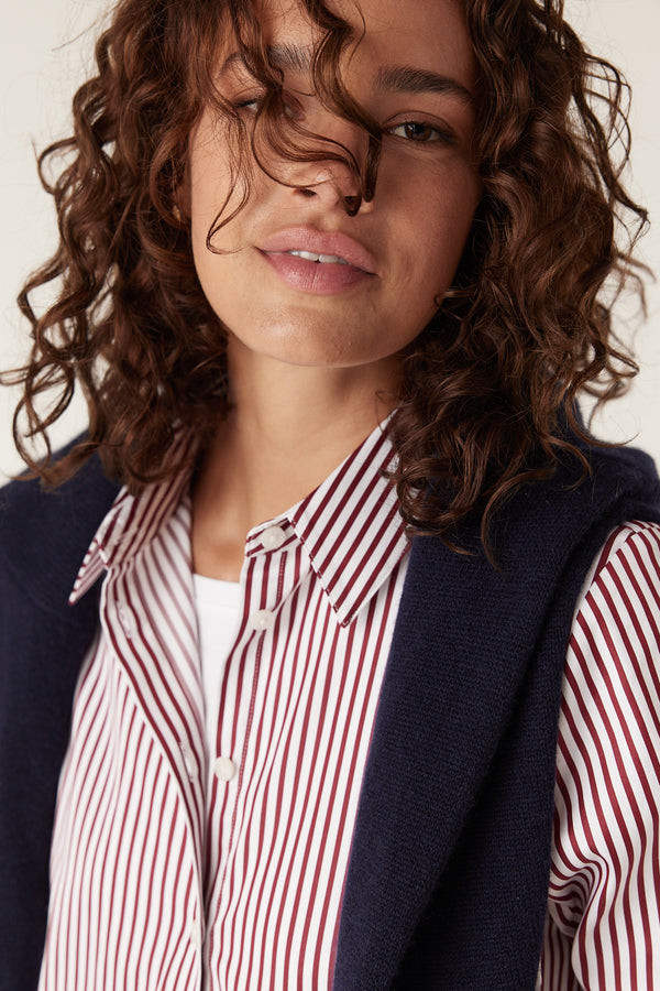 Woman wearing a white shirt with thin red stripes, a white undershirt, and a navy sweater draped over her shoulders.