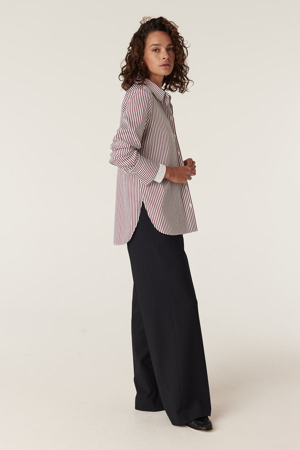 Woman wearing a red and white striped Henri shirt with long sleeves and a relaxed fit, paired with dark pants.