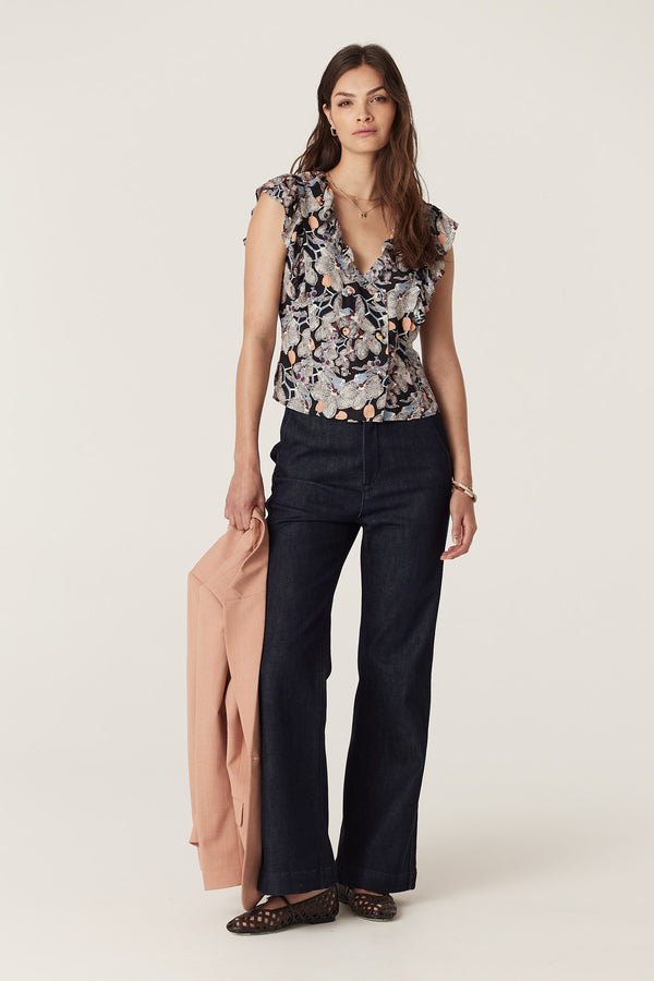 Woman in Leigh Frill Top, an orchid print silk blend V-neck top with frills, dark jeans, holding a peach jacket.