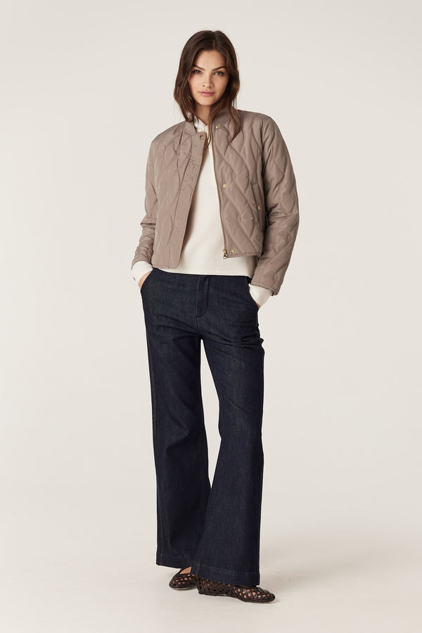 Woman in fawn Martina Puffer jacket with quilted cable design, white sweater, and dark jeans.