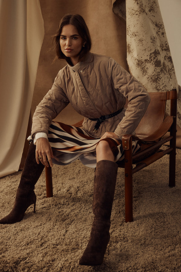 Woman in fawn quilted Martina Puffer jacket, belted, with a striped skirt and brown boots, sitting.