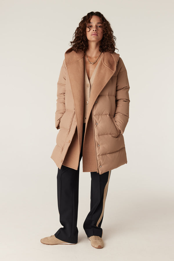 Woman wearing a camel Henderson Puffer coat with a wide collar, layered over a cardigan and black pants.