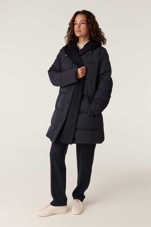 Woman wearing a navy Henderson puffer jacket with a ribbed collar and hood, over pinstripe pants.