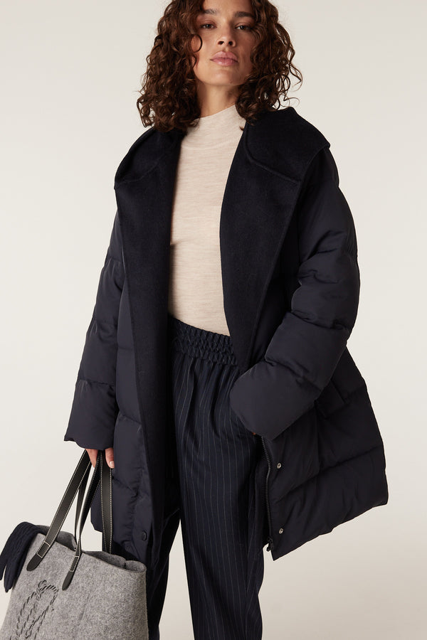 Woman in a navy Henderson Puffer jacket with a wide collar, beige top, pinstripe pants, and a grey tote bag.
