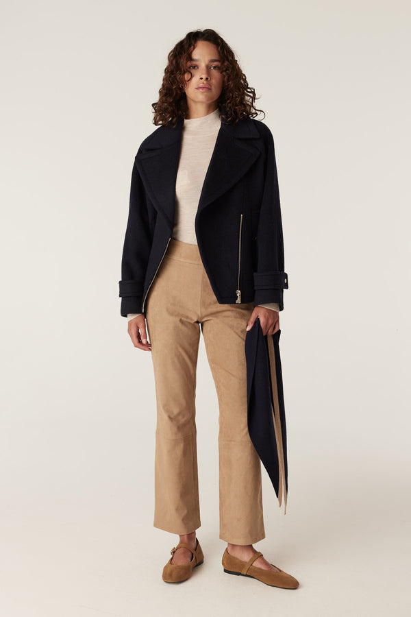 Woman in camel faux suede pants, cream turtleneck, and navy moto jacket with silver zipper.