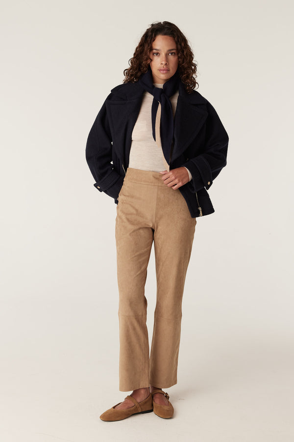Woman wearing camel Ines faux suede pants, a cream top, navy jacket, and navy neck scarf.