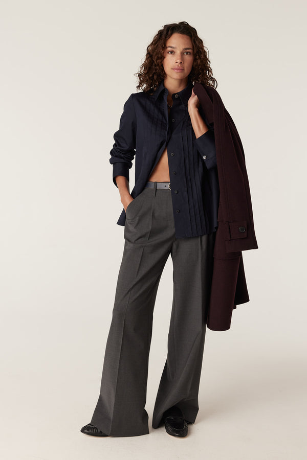 Woman in navy Chester Pintuck Shirt, grey wide-leg trousers, and a burgundy coat over her shoulder.