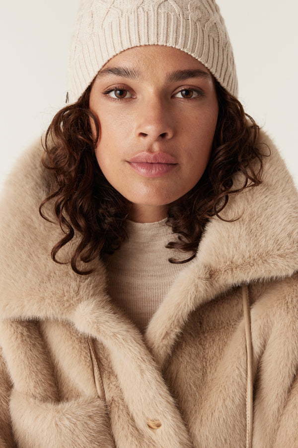 Close-up of a woman wearing a camel Franzi faux fur jacket with a wide collar and a cream knit beanie.