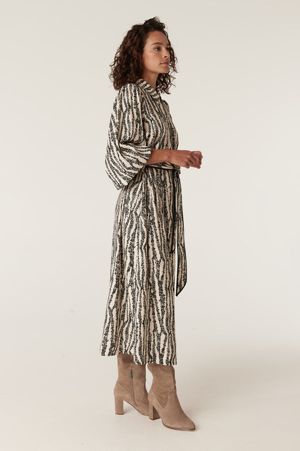 Woman in a long-sleeve Colette midi dress with a black speckle print, belted waist, and beige boots.