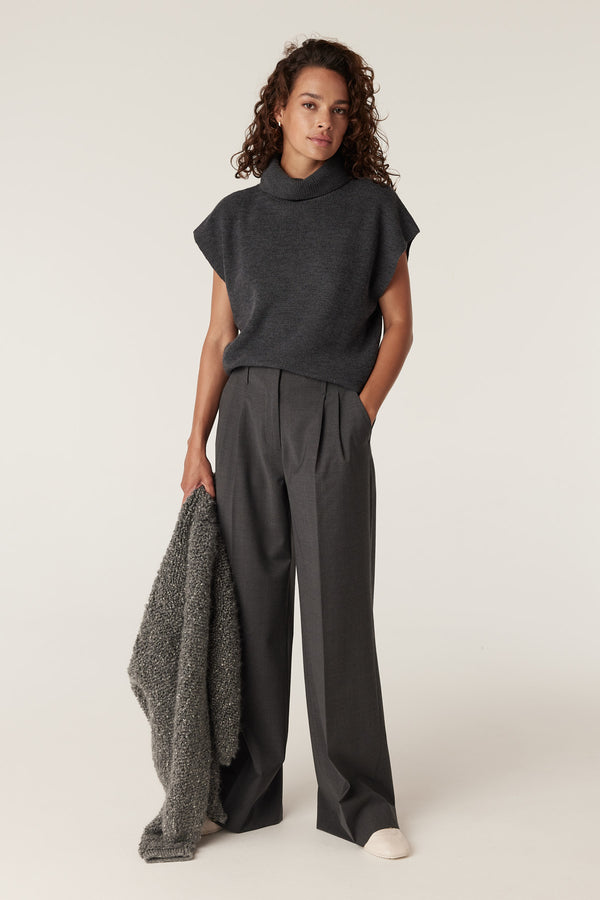 Woman wearing charcoal marle wool pants with pleats, a dark grey turtleneck vest, and holding a textured grey sweater.