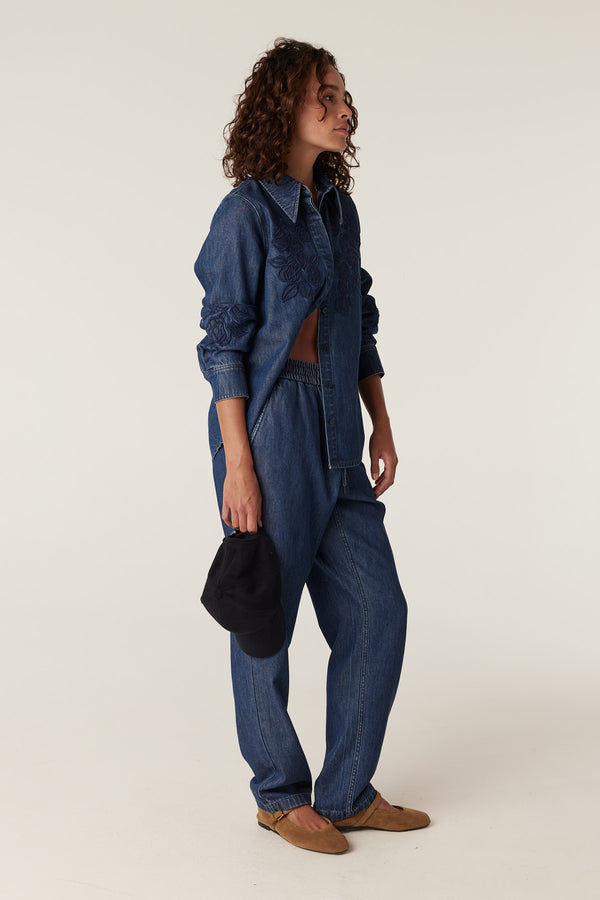 Woman in indigo denim jogger pants and matching embroidered shirt, holding a black cap.