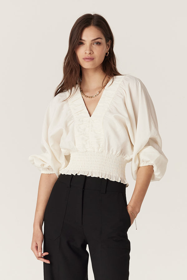 Model wears ecru Sasha Blouse with V-neck, ruched detail, batwing sleeves, and shirred waist.