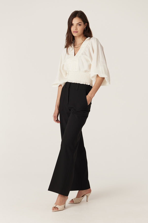 Model wears an ecru Sasha Blouse with V-neck, ruched detail, batwing sleeves, and shirred waist.