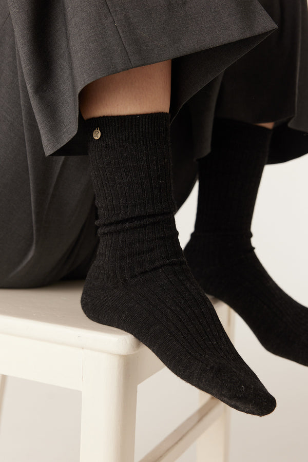 Charcoal ribbed merino wool socks on a person's feet, with a small gold logo on the ankle.