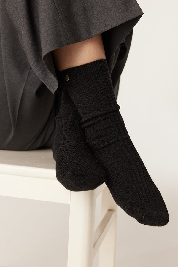 Charcoal ribbed merino wool socks with a subtle gold brand stud, worn by a person in dark pants.