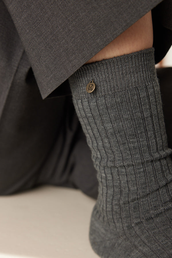 Mid-grey merino wool ribbed sock with a small bronze brand tag, worn with dark trousers.