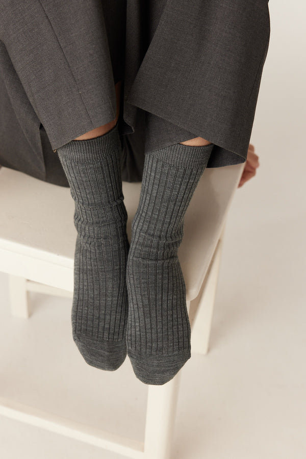 Mid-grey merino ribbed socks on feet, peeking from dark trousers, sitting on a white chair.