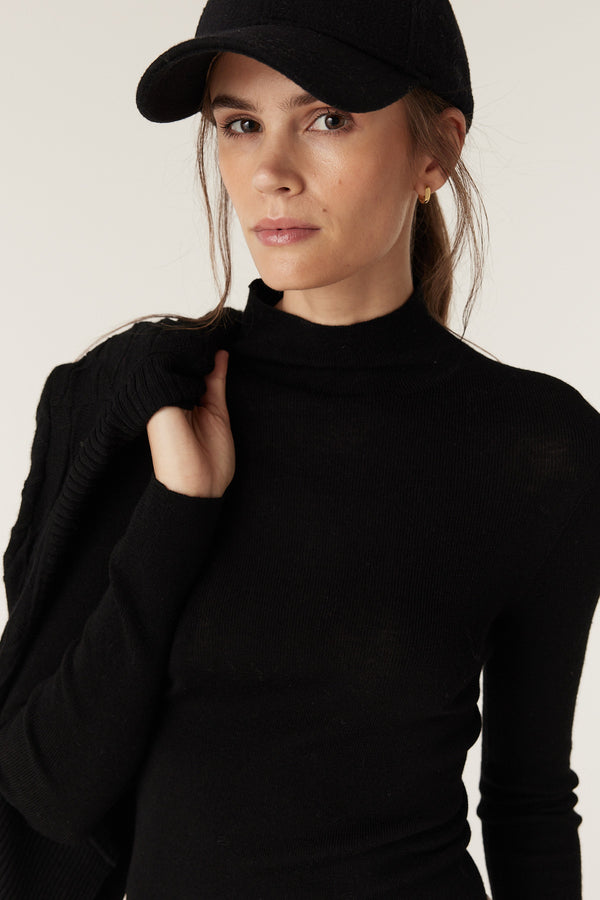 Woman in a black superfine merino wool turtleneck, wearing a black cap and gold earrings.