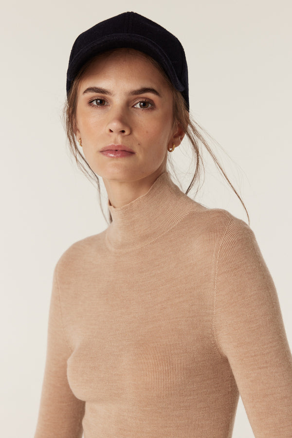 Woman in a latte superfine merino wool turtleneck with a mock neck and slim fit, wearing a navy cap.