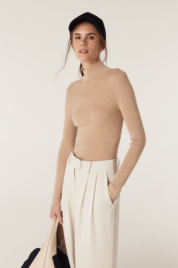 Model wears a slim-fit latte superfine merino wool turtleneck with cream pleated pants and a black cap.