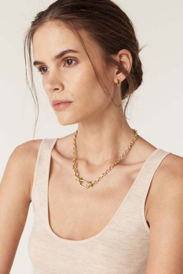 Woman in an oatmeal superfine merino wool singlet, wearing a gold chain necklace and stud earrings.