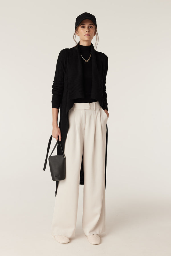 Woman wearing a black Merino wool longline cable knit cardigan, black top, cream wide-leg pants, and a black cap.